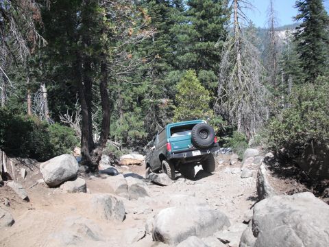 1991 Ford Bronco - 1991 Ford Bronco tackling a challenging off-road trail with large rocks and trees.