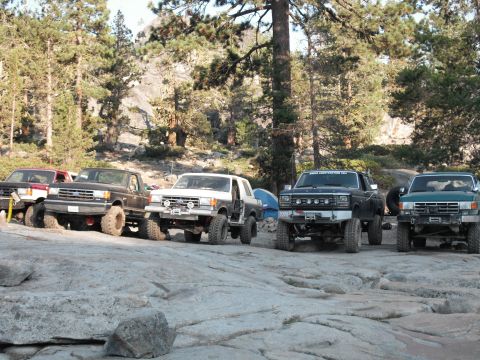 1991 Ford Bronco - A group of modified off-road vehicles are parked on a rocky terrain surrounded by trees.