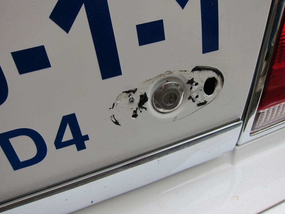 Close-up of a white car's rear with blue numbers and letters, showing a damaged lock cylinder