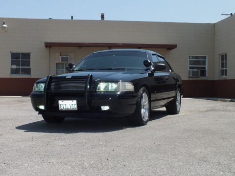 2003 Ford Crown Victoria - Lowered black sedan shows brush guard and headlights on, parked beside a tan building