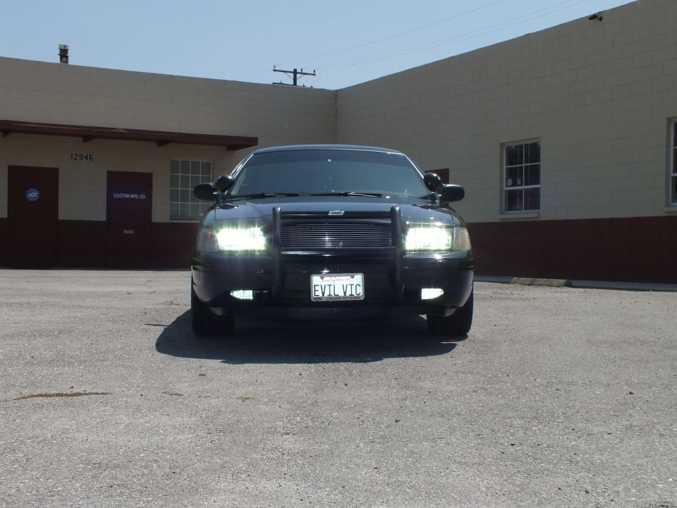 Head-on front view of black sedan with mesh grille, push bar and EVIL VIC plate, headlights lit