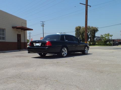 2003 Ford Crown Victoria - Rear-left angle of glossy black sedan, Ford oval on trunk, trees and stop sign behind
