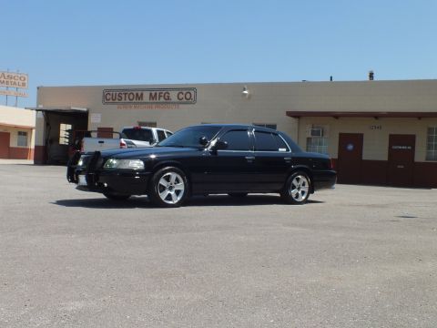2003 Ford Crown Victoria - Lowered black sedan with side spotlight and front guard parked before Custom Mfg Co; police interceptor sedan in background