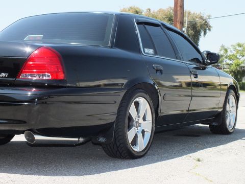 2003 Ford Crown Victoria - Black sedan rear quarter with SVT badge, Ford-branded mud flap and silver wheel near a tree