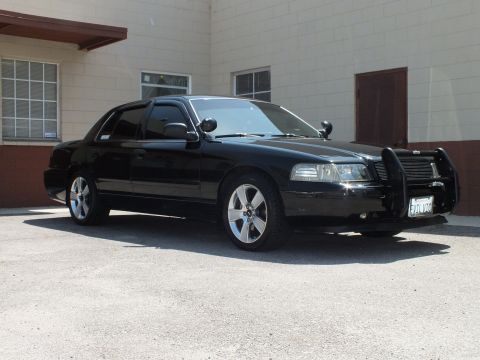 2003 Ford Crown Victoria - Lowered black sedan with police-style push bar and pillar spotlights parked beside a tan building