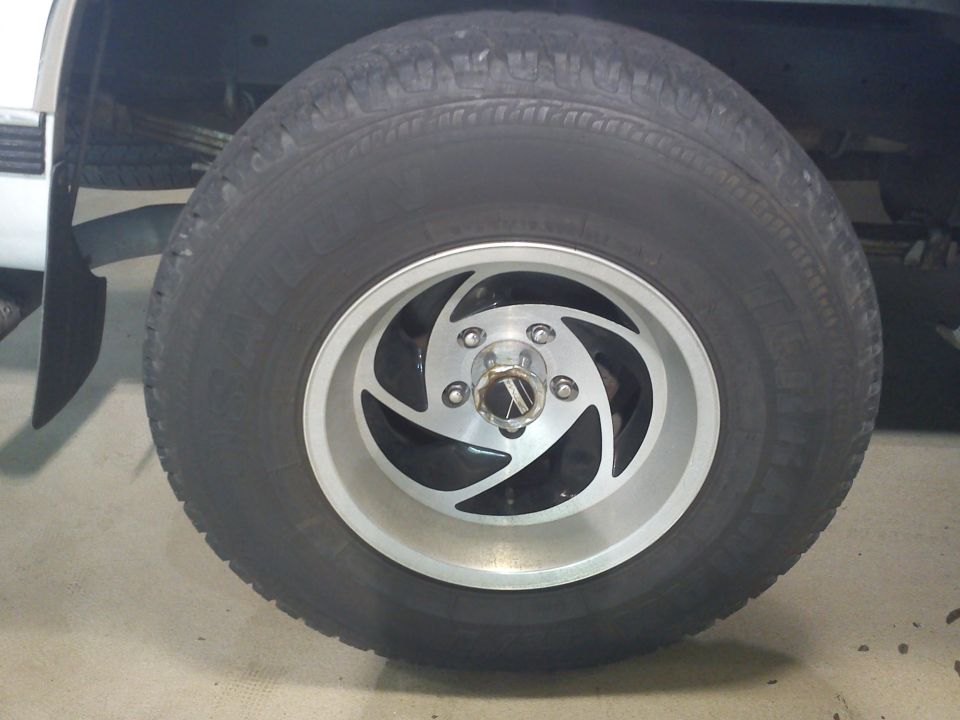 Close-up of a truck wheel with a silver and black swirl design rim and a tire. Supermotors.net logo visible