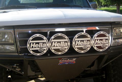 1996 Ford Bronco - The front grille of a white Ford truck is equipped with four Hella driving lights and aftermarket bumper stickers.