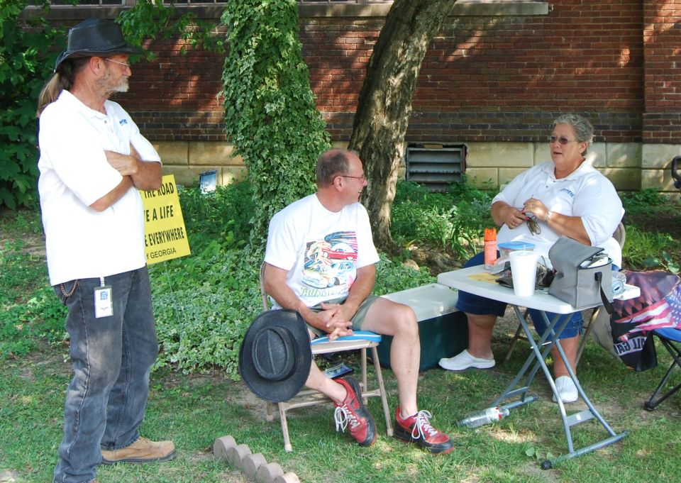 Three people are gathered outdoors, two sitting at a table and one standing. A yellow sign is visible behind the standing person