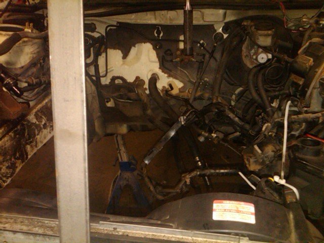 Engine bay with hoses, wires, and engine components visible. A jack stand supports part of the engine