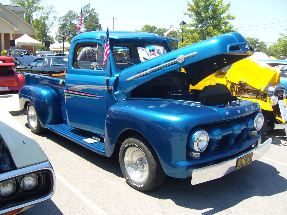 Blue 1951 Ford F-series pickup truck, hood open, front 3/4 view. American flag on driver's side window