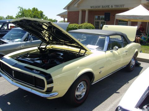 2007 Ford Mustang - A classic yellow Chevrolet Camaro RS convertible is displayed with its hood open, parked in front of a bank.