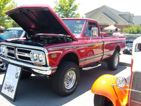 2007 Ford Mustang - A red 1972 GMC pickup truck is displayed with its hood open, parked next to an orange hot rod at a car show.