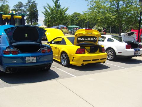2007 Ford Mustang - Three cars, a yellow Saleen Mustang, a blue Chevrolet Camaro, and a white Ford Mustang, are parked in a lot with their trunks open.