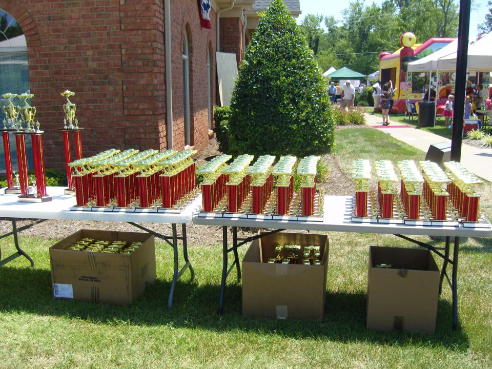 Tables full of red and gold trophies set up outside a brick building, with a bouncy house and tents in the background