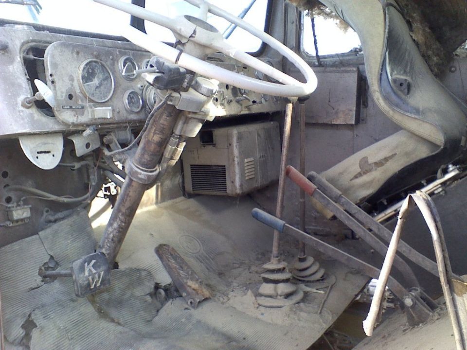 Dusty interior of an old truck cab. Shows steering wheel, dashboard with gauges, pedals, and gear shifters