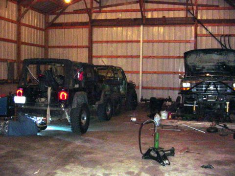 1992 Ford Bronco - A person works on the underside of a black Jeep Wrangler inside a garage, with a Ford truck visible in the background.