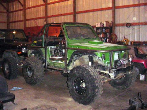 1992 Ford Bronco - A heavily modified green vehicle with a roll cage and exposed interior sits in a garage next to another vehicle.