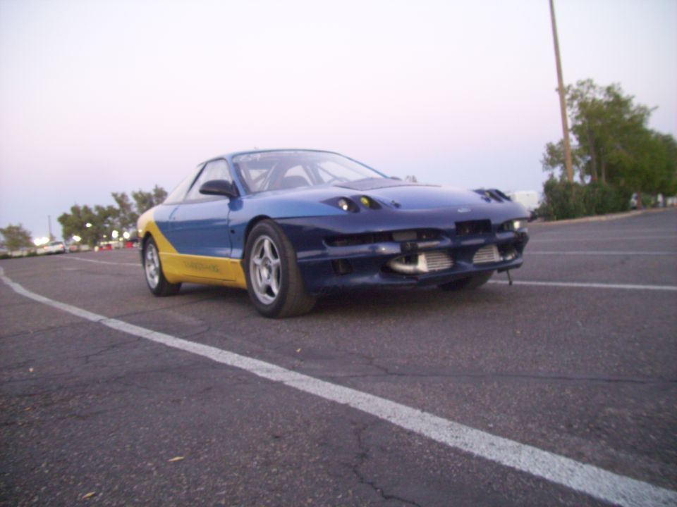 1995 Ford Probe with blue and yellow paint scheme, aftermarket front bumper, and roll cage visible through windows