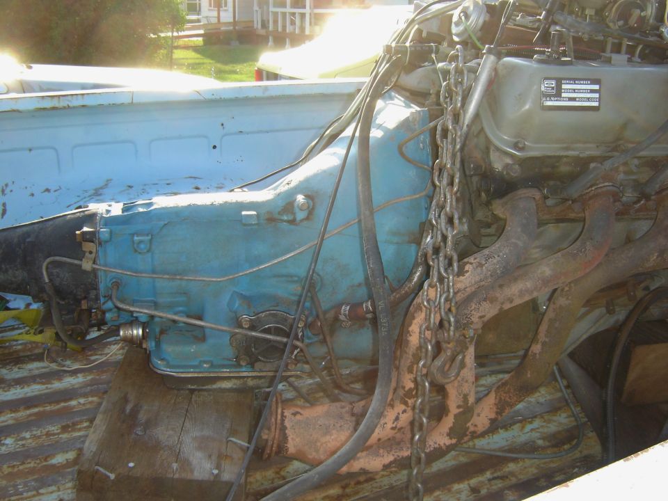 Engine and transmission assembly, blue and rusty, with chains attached, in the bed of a light blue truck