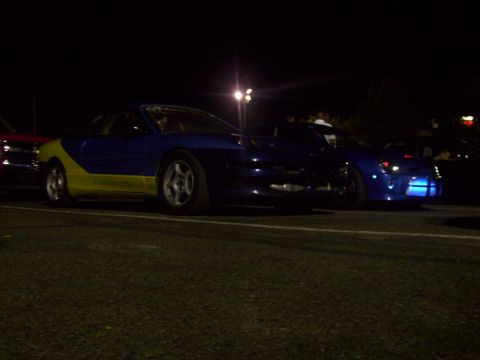 1995 Ford Probe - A blue and yellow sports car is parked at night next to another blue sports car.