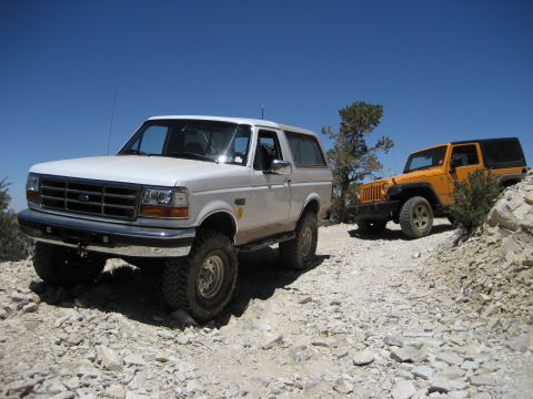 1996 Ford Bronco - Two off-road vehicles, a 1996 Ford Bronco and an orange Jeep, navigate a rocky path under a clear blue sky.