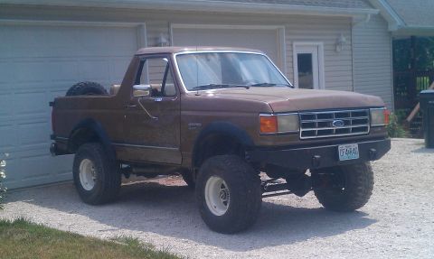 1987 Ford Bronco - A brown 1987 Ford Bronco is parked in front of a garage, modified with a lift kit and large tires.
