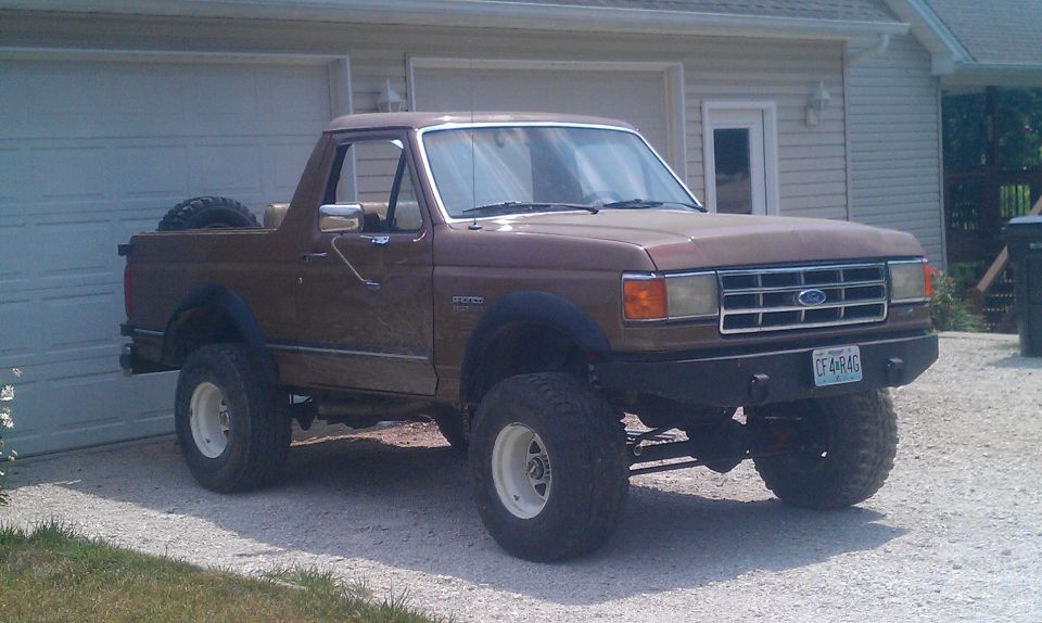 Brown 1987 Ford Bronco with lifted suspension and off-road tires parked in front of a garage