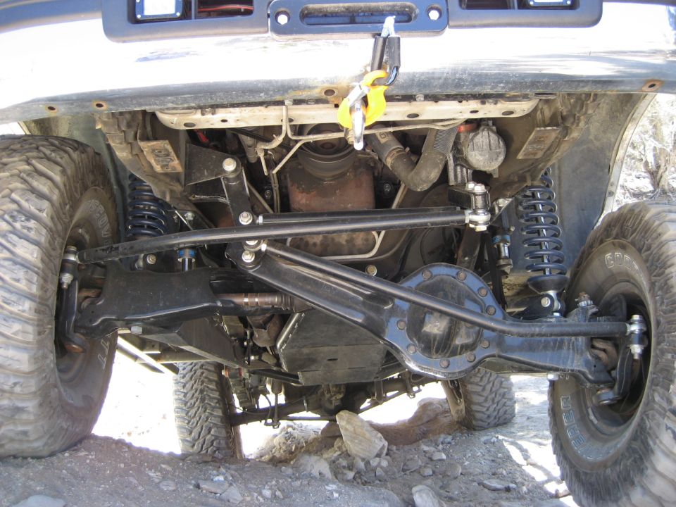 Undercarriage of a lifted 4x4 with large tires, showing suspension and drivetrain components. Winch hook visible on front