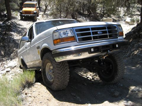 1996 Ford Bronco - Two off-road vehicles, a white 1996 Ford Bronco and a yellow Jeep, navigate a rocky trail in a wooded area.