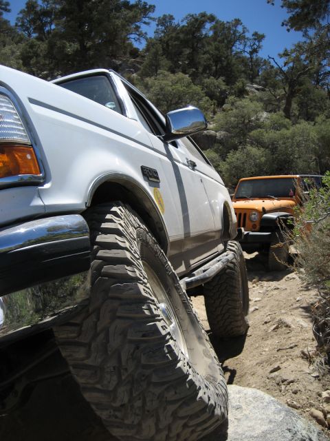 1996 Ford Bronco - Two off-road vehicles navigate a narrow, rocky path through a wooded, mountainous area.