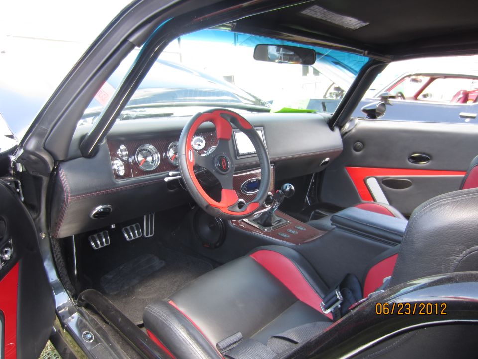 Interior view of a car with black and red seats, custom steering wheel, and dashboard. Another car is visible through the window
