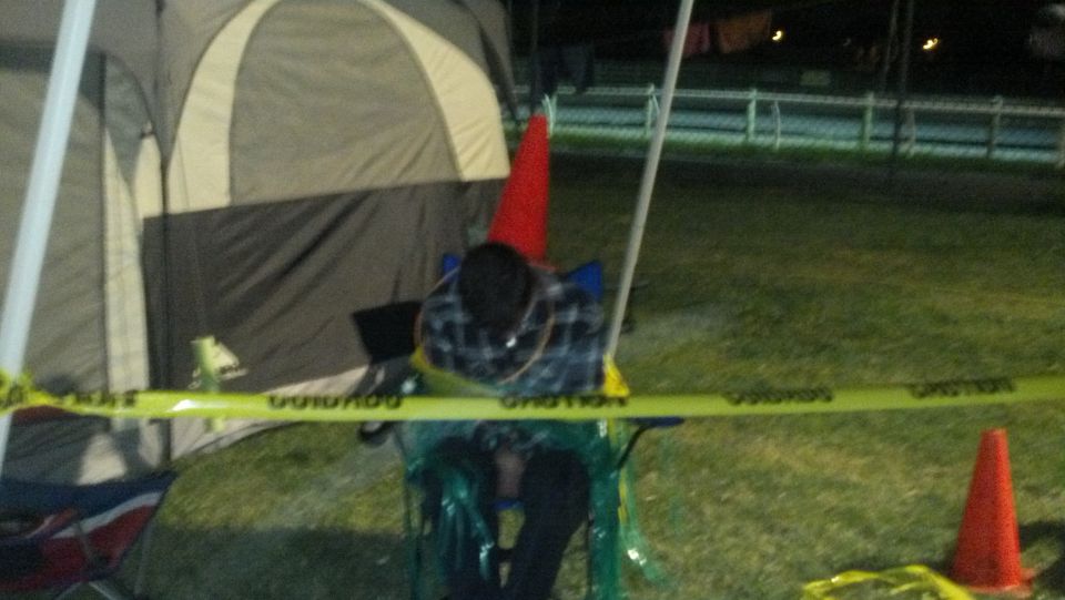 Person sitting in a chair, wrapped in caution tape, next to a tent. Orange cones are visible