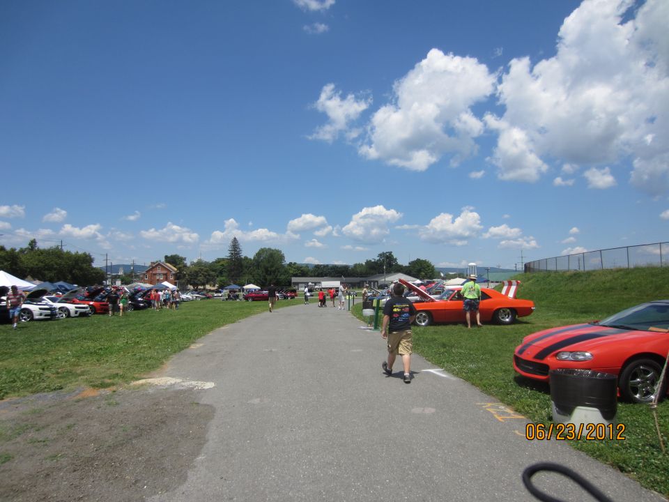 Orange muscle car with black racing stripes and white accents on the rear, parked on grass at an outdoor car show