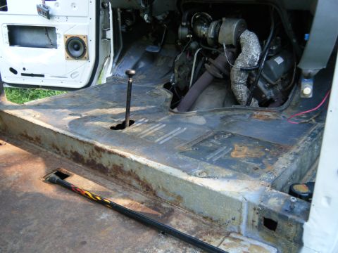 1988 Ford E350 - The interior floor of a van shows significant rust and exposed engine parts, with a speaker mounted in the door panel.