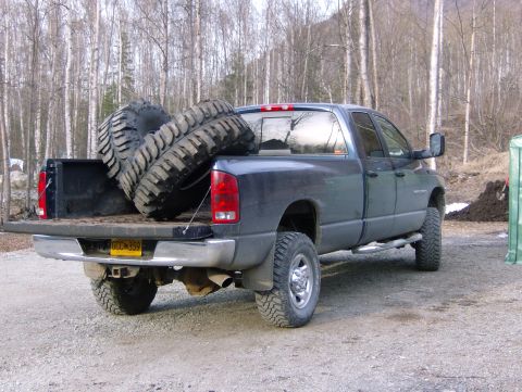1983 Chevrolet C/K Pickup - A dark-colored pickup truck is parked outdoors with its tailgate down, carrying two oversized tires in the bed.