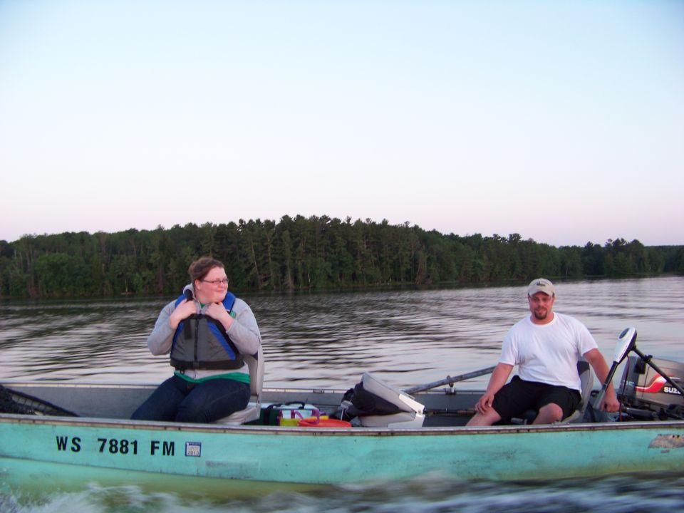 Two people in a light blue boat on a lake with trees in the background. Boat has registration WS 7881 FM