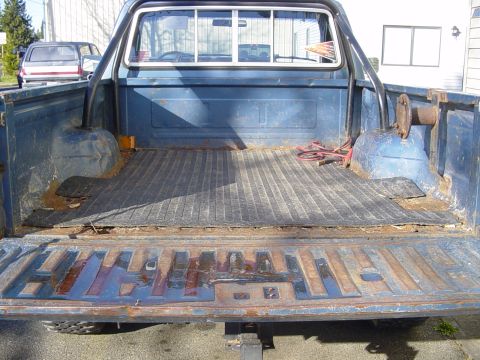 1987 Ford Bronco - The bed of a blue pickup truck shows signs of wear and tear, with a rubber mat and rusty tailgate.