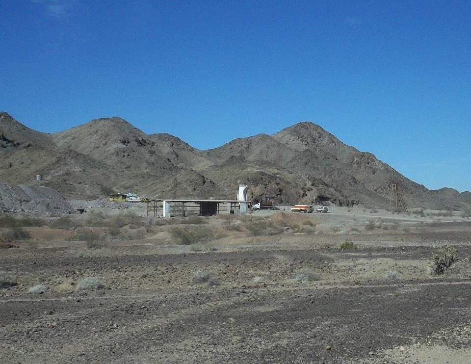 Desert landscape with mountains, a building complex, and several vehicles including trucks and what appears to be a bus