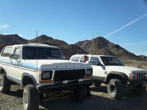1979 Ford Bronco - Two off-road vehicles, a 1979 Ford Bronco and a Jeep Cherokee, are parked in a dry, mountainous area under a clear blue sky.