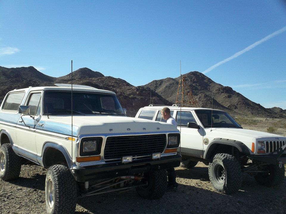 White and blue 1979 Ford Bronco with lifted suspension, parked next to a white Jeep Cherokee in a desert landscape