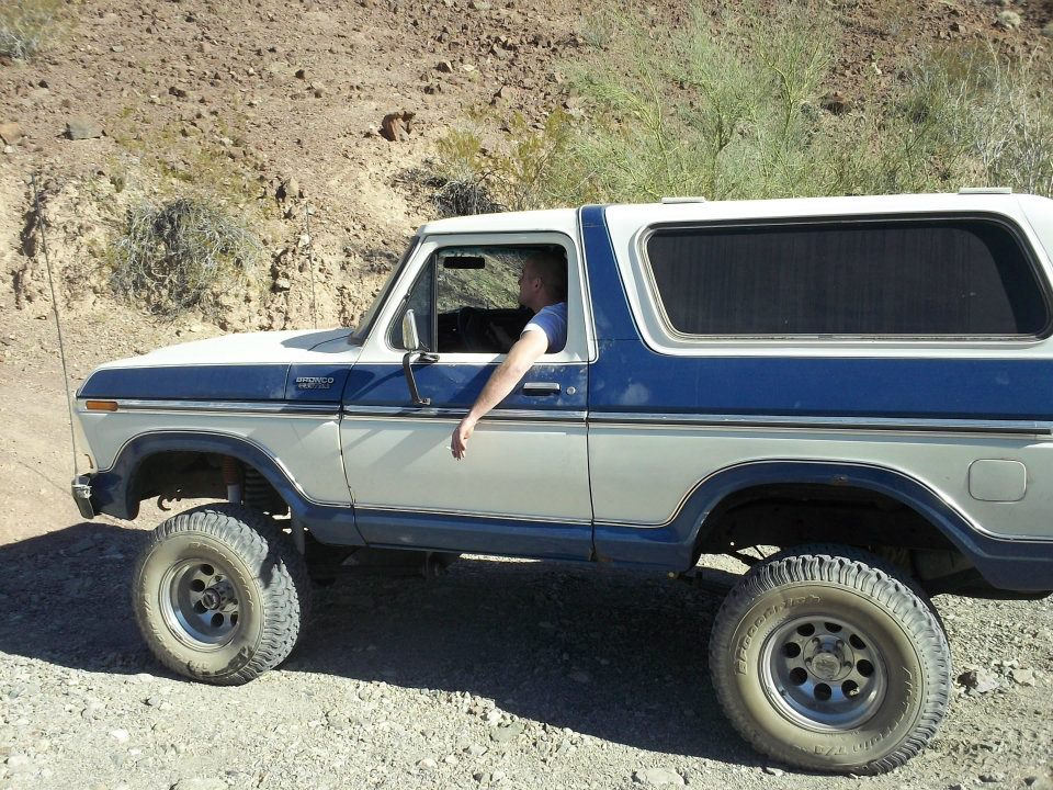 Side view of a blue and white 1979 Ford Bronco driving on a dirt road