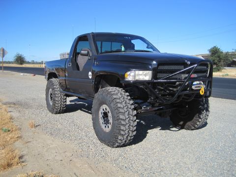 1989 Ford Bronco - A modified black Dodge Ram pickup truck sits parked on a gravel shoulder under a clear blue sky.