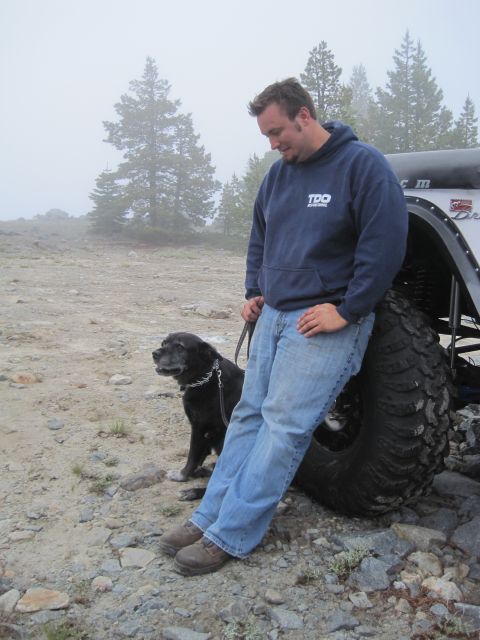 1989 Ford Bronco - A man and his dog take a break next to a large tire, with a foggy, tree-covered landscape behind them.