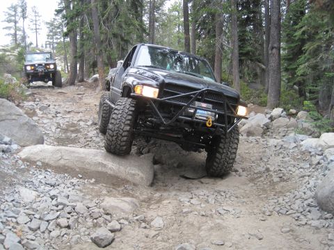 1989 Ford Bronco - A black pickup truck drives off-road on a rocky path, with another vehicle visible in the background.