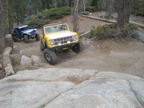 1989 Ford Bronco - Two modified off-road vehicles navigate a challenging dirt and rock path in a wooded area.