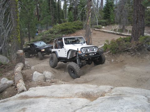 1989 Ford Bronco - Two off-road vehicles navigate a challenging dirt path surrounded by trees and rocks.
