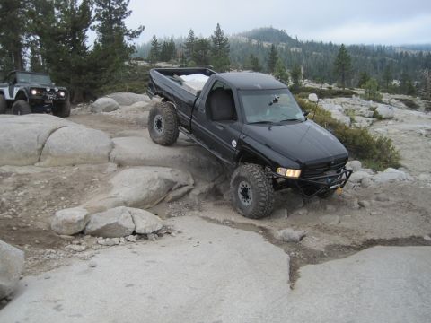 1989 Ford Bronco - A black pickup truck maneuvers over large rocks on an off-road trail, with another vehicle visible in the background.