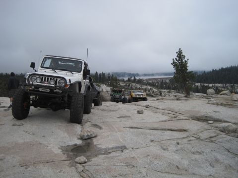 1989 Ford Bronco - Multiple off-road vehicles are parked on a rocky surface under a cloudy sky.