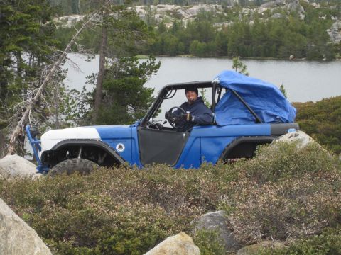 1989 Ford Bronco - A man sits in the driver's seat of a modified off-road vehicle, surrounded by bushes and rocks, with a lake in the background.