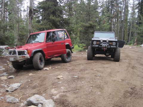 1989 Ford Bronco - Two off-road SUVs are parked on a dirt path surrounded by trees, with one vehicle's door open.